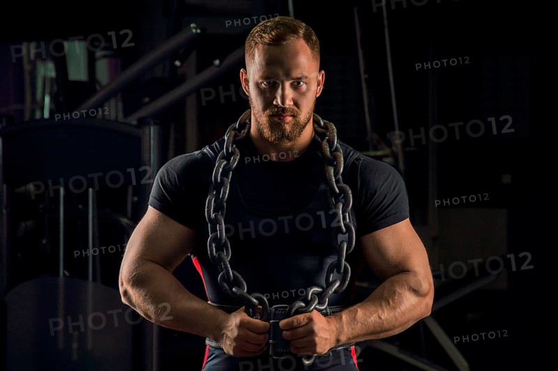 Handsome athlete with a chain around his neck stands in the gym and looks straight ahead