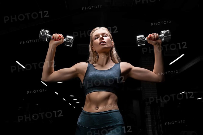 Charming blonde posing in the gym with dumbbells in her hands. View from below. The concept of sports