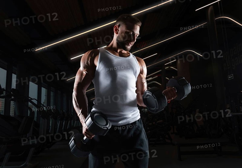 Muscular man in a white t-shirt works out in the gym with dumbbells. Biceps pumping. Fitness and bodybuilding concept.