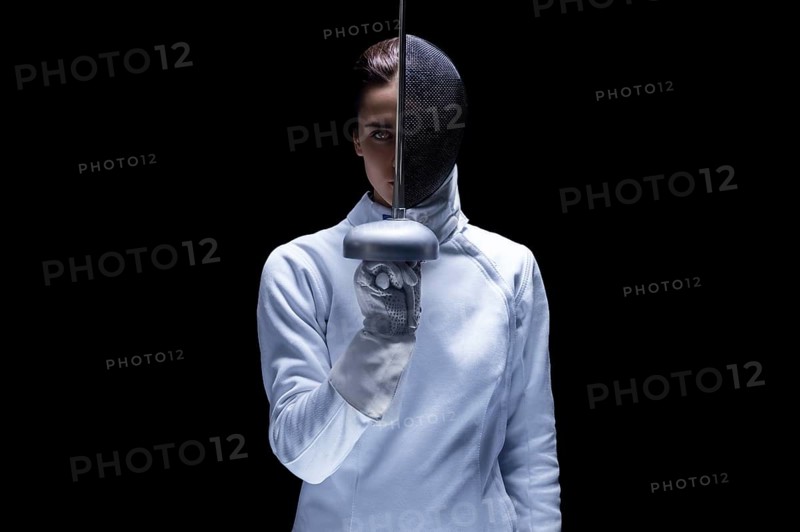 Charming girl in a fencer costume posing on a black background. The sword separates the head into a face and a mask. The concept of fencing.
