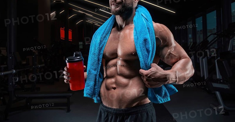 Portrait of a muscular guy with a naked torso and abs. Gym. Blue towel and red shaker. Fitness concept
