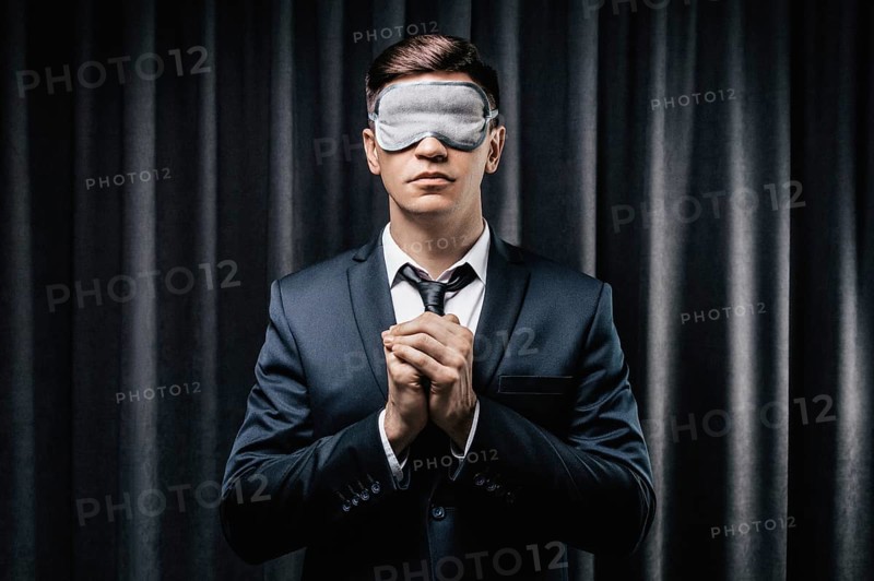 Portrait of a man in a mask for sleeping. He prays against a black curtain. The concept of blind dates