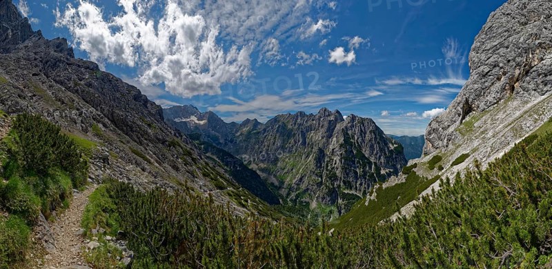 A narrow mountain path winds its way through a landscape of lush greenery and rock faces under a cloudy sky
