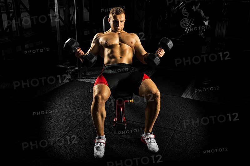 Professional athlete makes an exercise on the biceps by lifting dumbbells while sitting on the bench. View from above