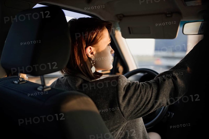 Young beautiful smiling woman taxi driver in a jacket sits behind the wheel of a car and adjusts the distant view mirror