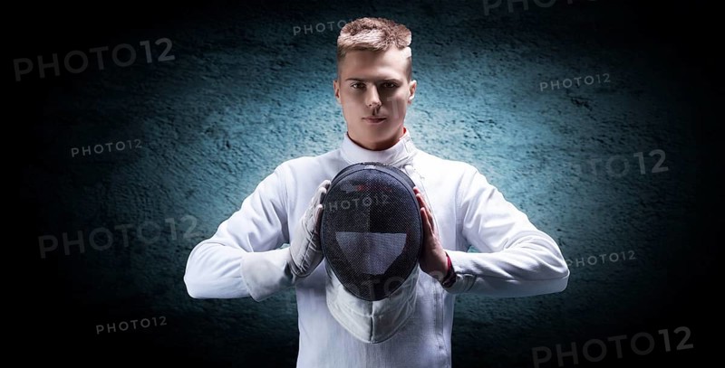 Portrait of a male fencer against a blue spot on the wall. He holds a helmet in front of him. The concept of fencing.