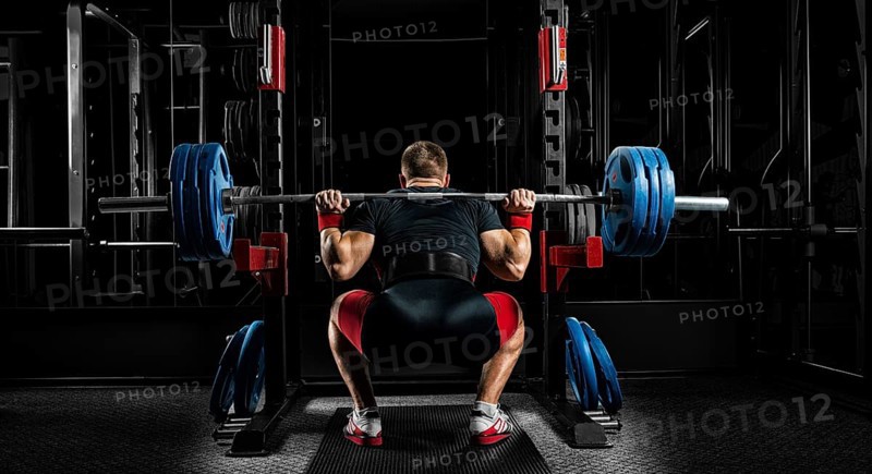 Professional athlete sits with a barbell on his shoulders and prepares to stand with her. View from the back