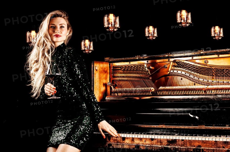 Charming girl in a green evening dress posing with a glass of wine in her hand near the retro piano.