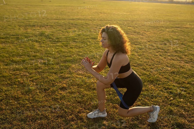 Young curly athletic girl in sportswear performs lunges with resistance band outdoors on the grass during sunset