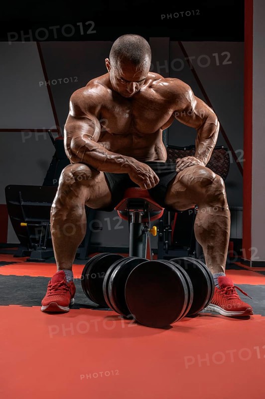 Muscular man sits on a bench near huge dumbbells. Bodybuilding and powerlifting concept.