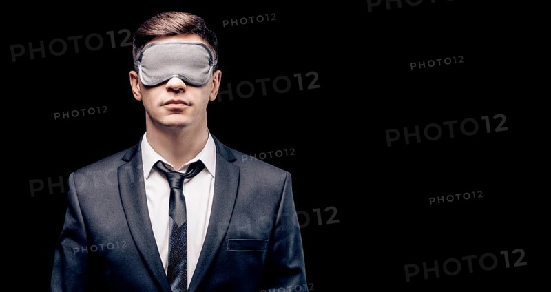 Portrait of a man in a mask for sleeping. He stands against a black wall. Blind business concept. Work during the crisis.