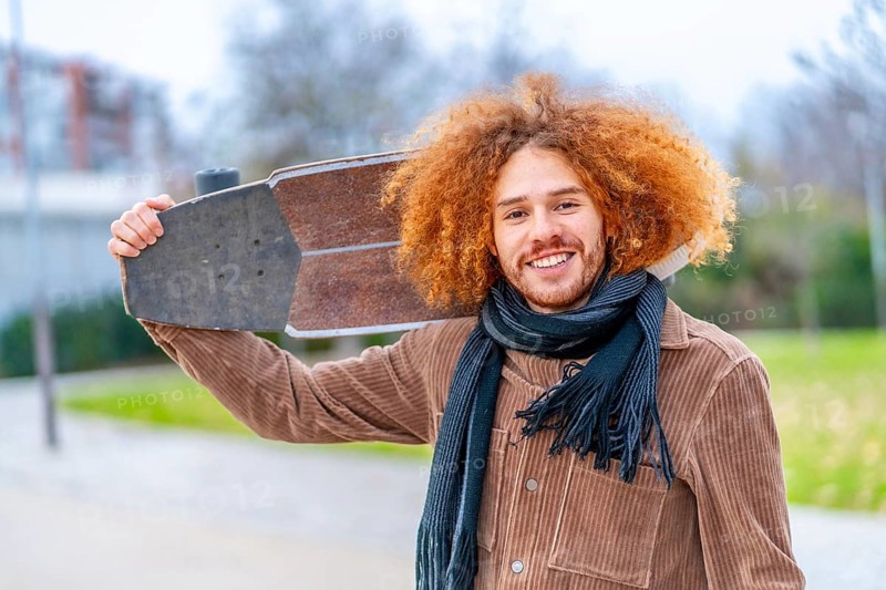 Portrait of a redheaded skater carrying a skate and smiling at camera in an urban park