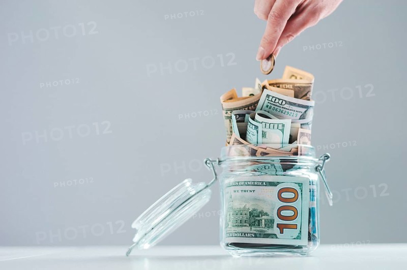 Female hand puts a coin into a glass piggy bank full of money. Thrift concept.