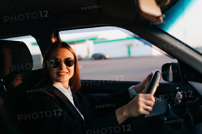 A young beautiful stylish girl driver in a jacket and sunglasses behind the wheel of a car greets passengers