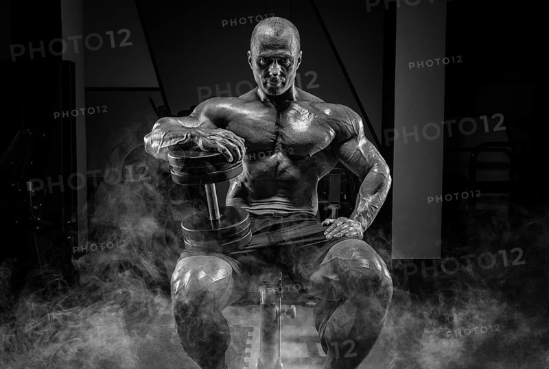 Muscular man sits on a bench in smoke with dumbbells. Bodybuilding and powerlifting concept.