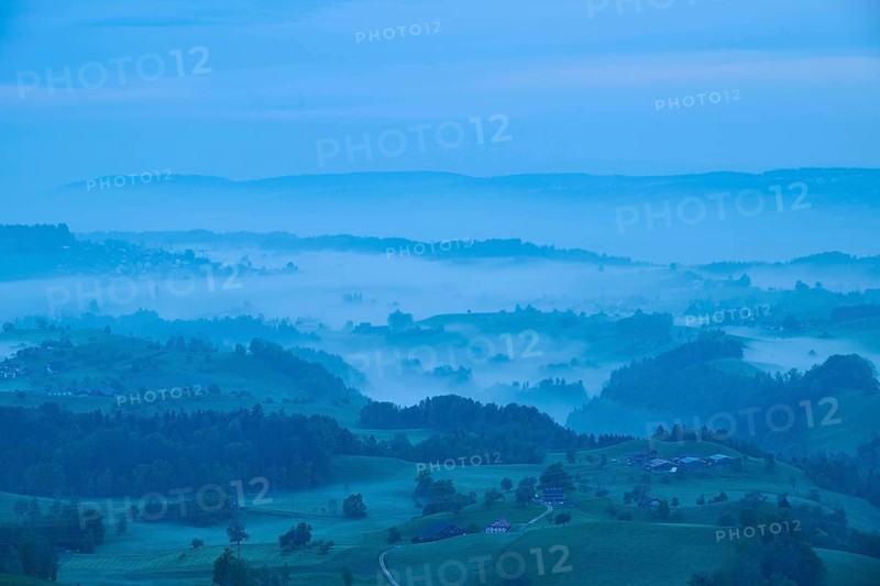 Moraine hill landscape with meadows and trees and some farms with morning fog and cloudy sky