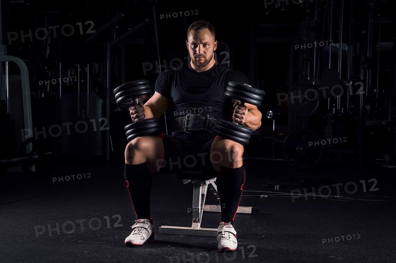 Impressive athlete sits on a bench in the gym with two dumbbells on his legs