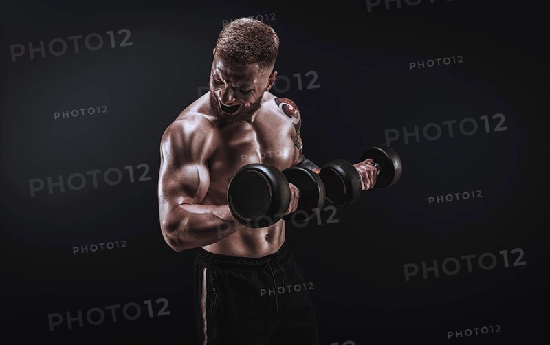 Young muscular guy pumping biceps with dumbbells. Concentrated flexion. Fitness and nutrition concept.
