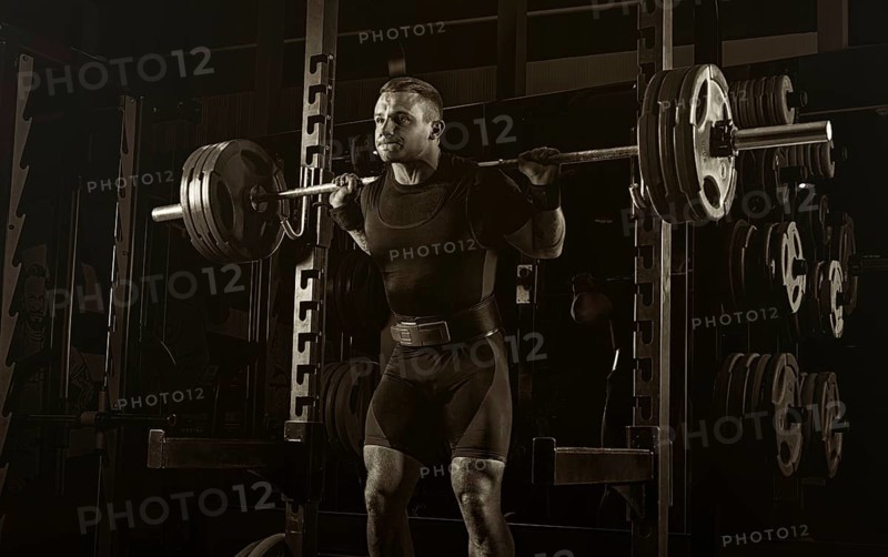 The weightlifter stands with a very heavy barbell on his shoulders. He is at the top and is preparing to sit down