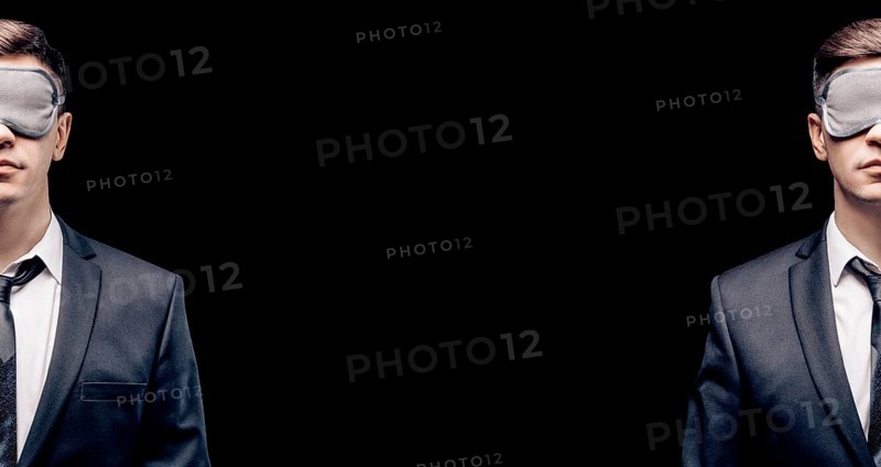 Portrait of a man cut in half in a sleep mask. He is standing on a black background. Concept of blind business