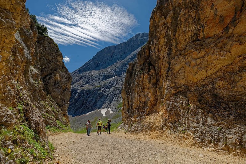 A group of hikers on a mountain path surrounded by imposing rocks under a blue sky with foehn clouds