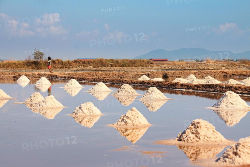 Salt piles symmetrically reflected in the water under a clear blue sky with a person in the distance. Kampot