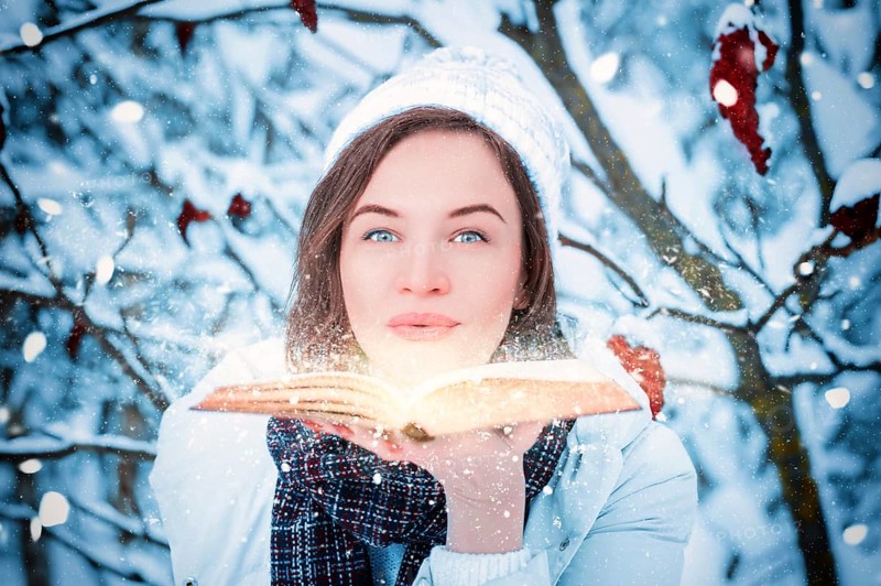 Charming woman blows bright dust from the pages of a book in the winter forest. Fairy tale concept. Christmas. New Year's holidays.