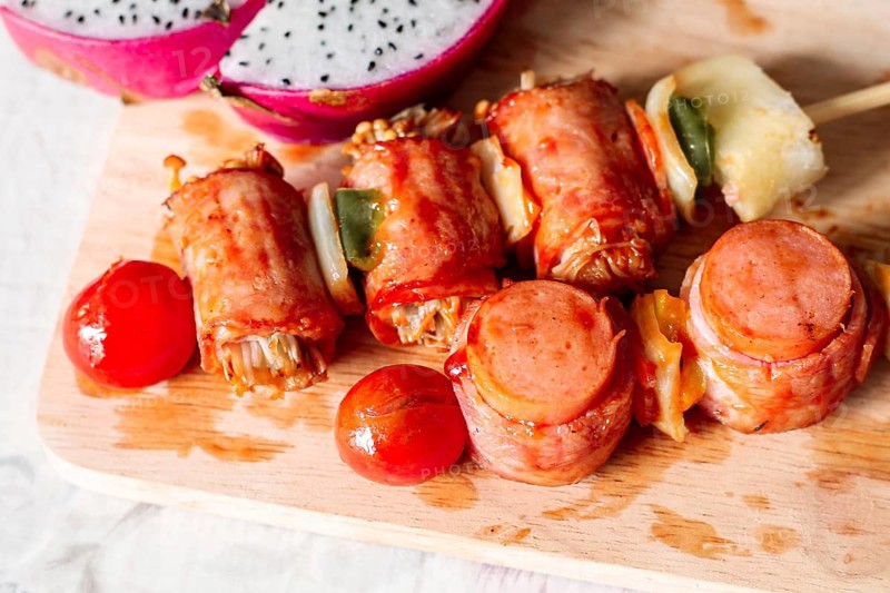 Skewers with bacon-wrapped sausages and vegetables on a wooden cutting board with dragon fruit