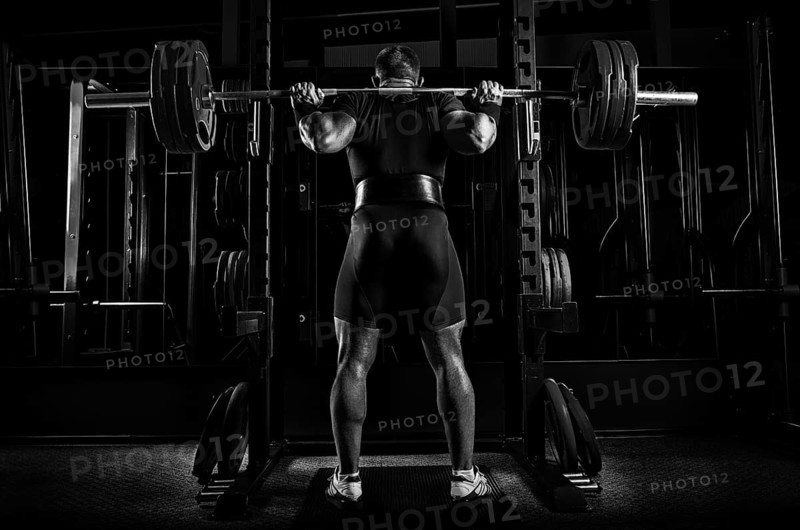 Professional athlete is standing with a barbell on his shoulders and is about to sit down with her. View from the back