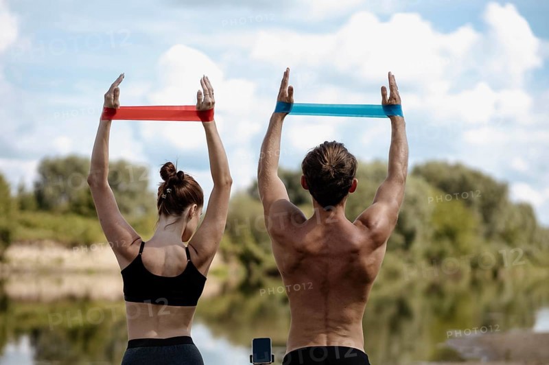 Young athletic couple doing exercise with resistance band on the shore of a lake on a sunny day