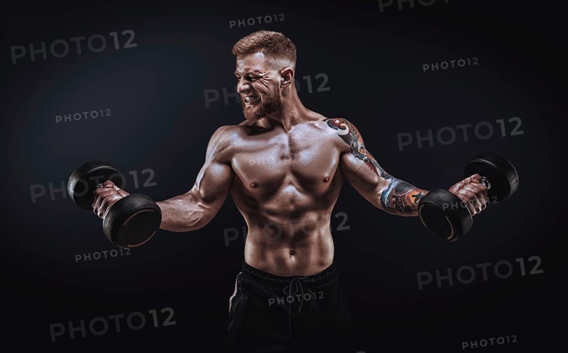 Young muscular guy pumping biceps with dumbbells. Concentrated flexion. Fitness and nutrition concept.