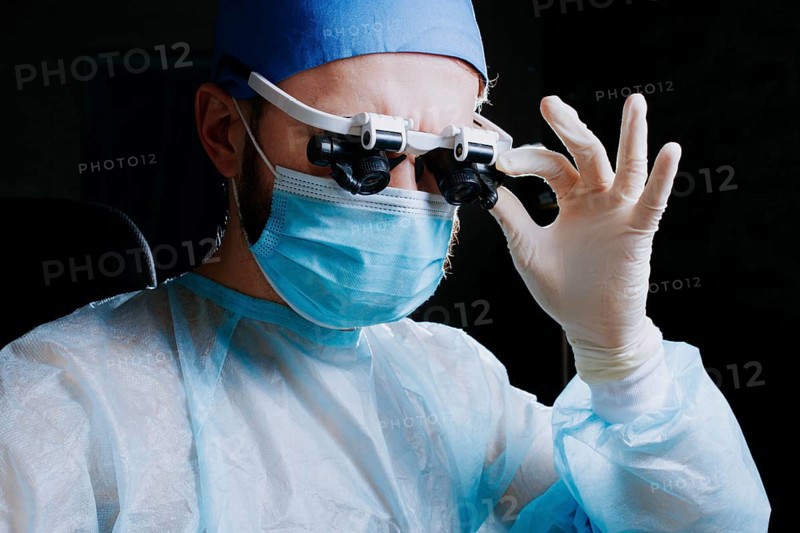 Surgeon wearing binocular magnifying glasses operates on a patient in a dark operating room