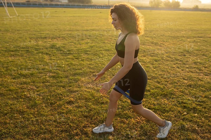 Young curly athletic girl in sportswear performs lunges with resistance band outdoors on the grass during sunset