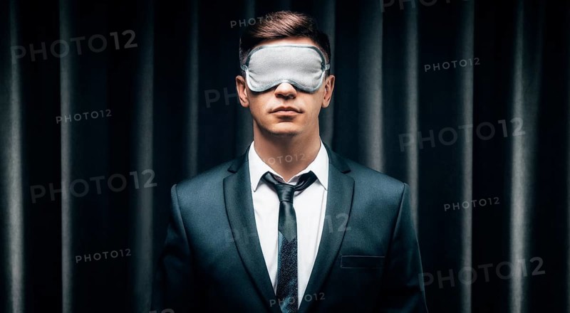 Portrait of a man in a mask for sleeping. He stands against the backdrop of a curtain. The concept of blind dates