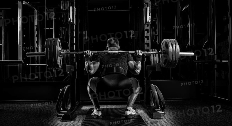 Professional athlete sits with a barbell on his shoulders and prepares to stand with her. View from the back