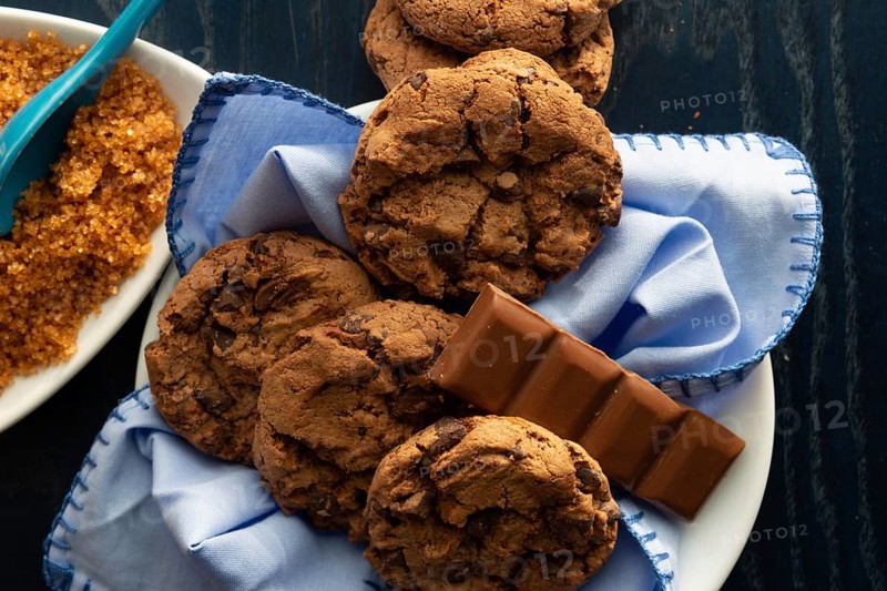 Chocolate chip cookies on a white plate beside brown sugar and a piece of milk chocolate
