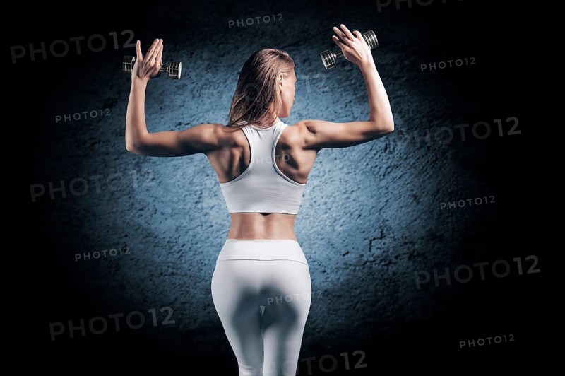 Adorable girl is training in the gym. She lifts the dumbbells up. Back view. The concept of bodybuilding