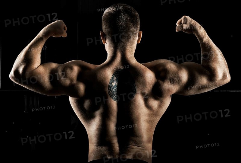 Huge weightlifter is standing with his back to the camera and straining his hands demonstrating a double bicep. Back view