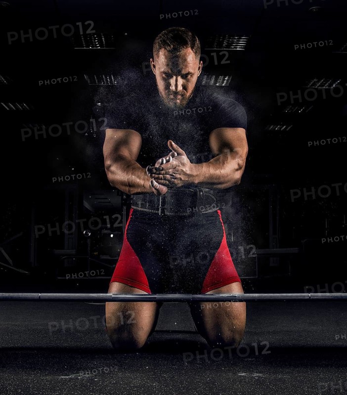 Ahlete kneels in the gym before the bar and claps his hands after which the dust from the chalk flies
