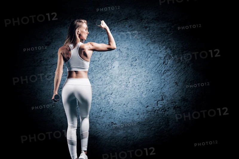 Adorable girl is training in the gym. She lifts the dumbbells up. Back view. The concept of bodybuilding