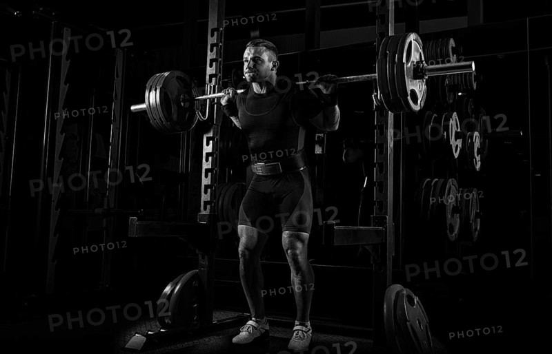 The weightlifter stands with a very heavy barbell on his shoulders. He is at the top and is preparing to sit down