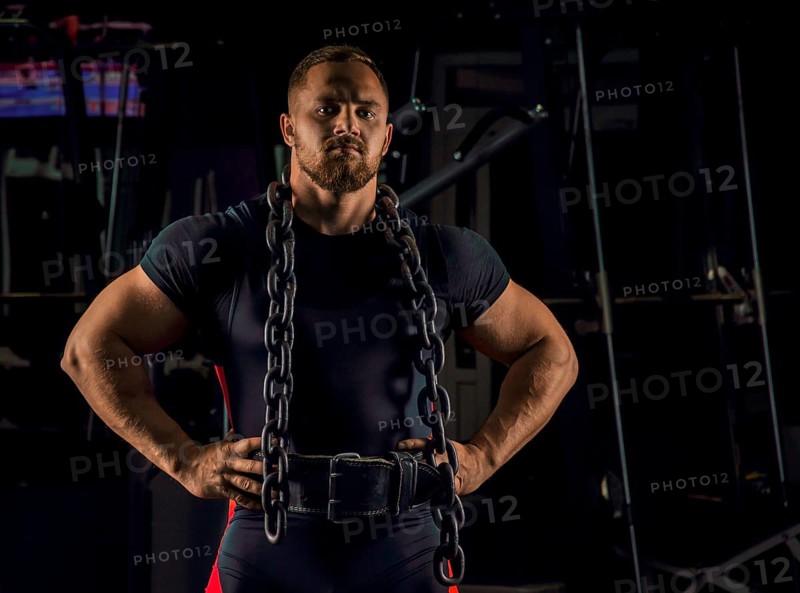 Handsome athlete with a chain around his neck stands in the gym and looks straight ahead