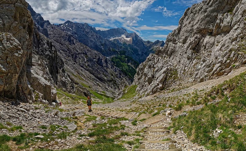 A mountaineer walks down a stony path in a mountainous valley under a cloudy sky