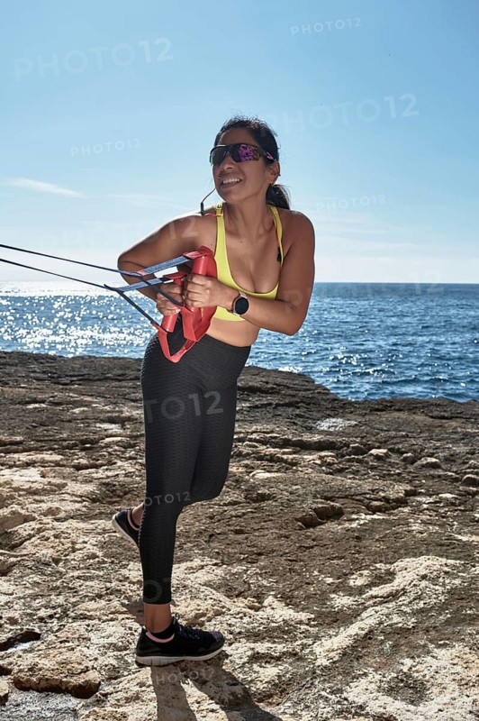 A woman in sporty attire exercises with a resistance band by the sunny seaside