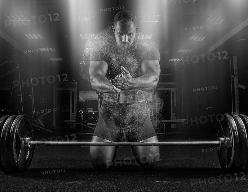 Ahlete kneels in the gym before the bar and claps his hands after which the dust from the chalk flies