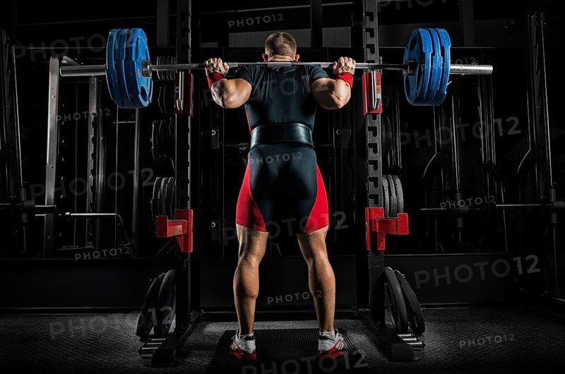 Professional athlete is standing with a barbell on his shoulders and is about to sit down with her. View from the back