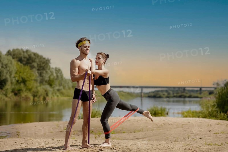Young athletic couple doing exercise with resistance band on the shore of a lake on a sunny day