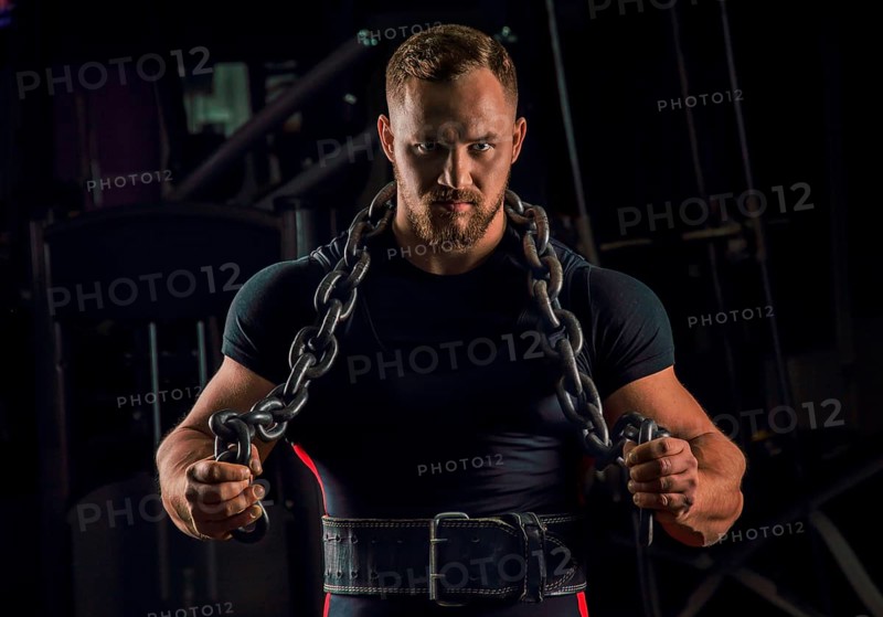 Handsome athlete with a chain around his neck stands in the gym and looks straight ahead