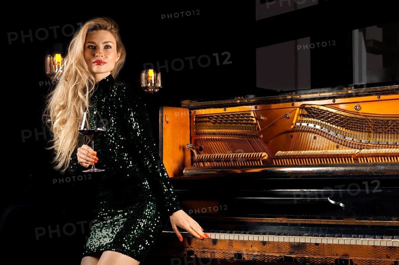 Charming girl in a green evening dress is smiling and holding a glass of wine in her hand while sitting on a retro piano.