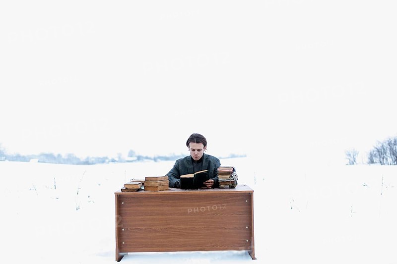 A young handsome intelligent man in a jacket sits at a table in a field and reads ancient books in wintera young handsome intelligent man in a jacket sits at a table in a field and reads ancient books in winter